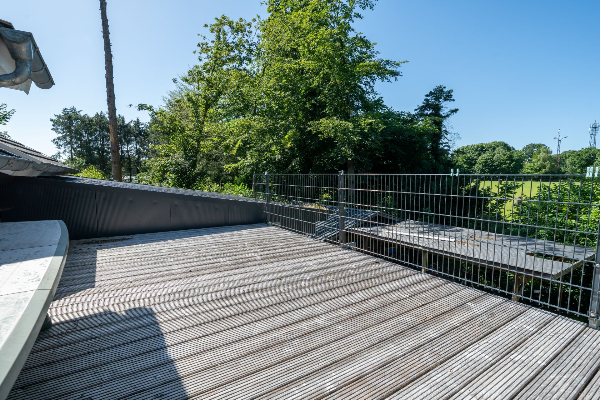 Große Holzterrasse mit Gitterzaun, dahinter Bäume und viel Grün im Hintergrund.