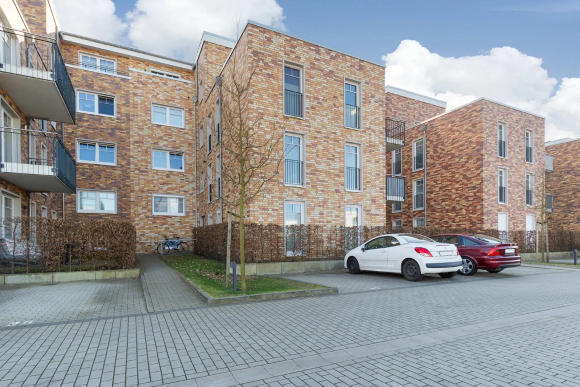 Mehrstöckiges Wohnhaus aus Klinker mit parkenden Autos und Fahrrad vor dem Gebäude.