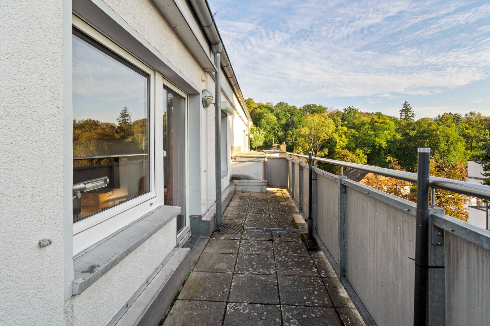 Langer Balkon mit Geländer, Blick auf Bäume und Fensterfront eines Wohngebäudes.