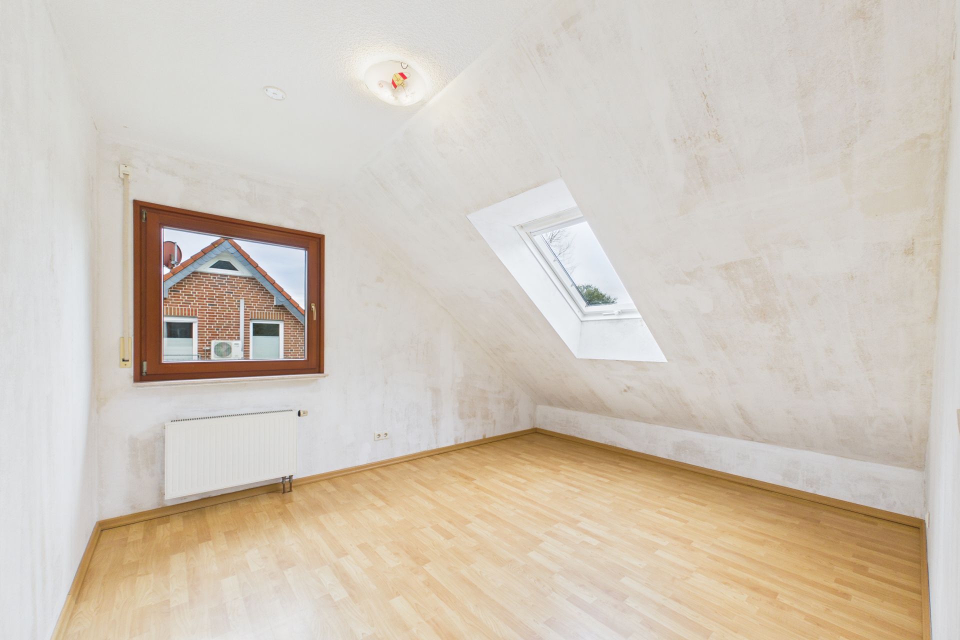 Helles leeres Zimmer mit Schrägdecke, Holzfußboden und zwei Fenstern.