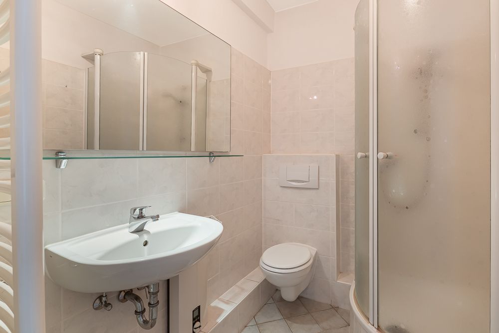 Kleines Bad mit Waschbecken, WC, Spiegel und abgerundeter Duschkabine.