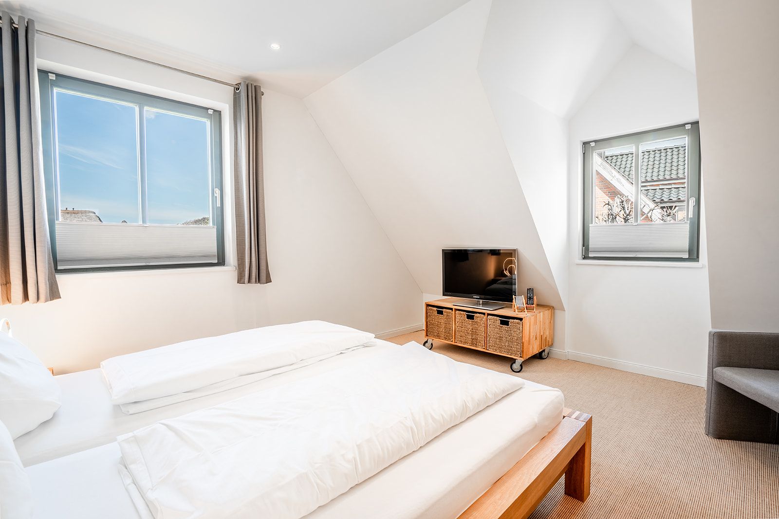 Helles Schlafzimmer mit Doppelbett, zwei Fenstern, Fernseher und Kommode aus Holz.