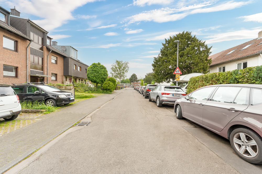 Ruhige Wohnstraße mit parkenden Autos, Häusern und Bäumen an beiden Seiten.