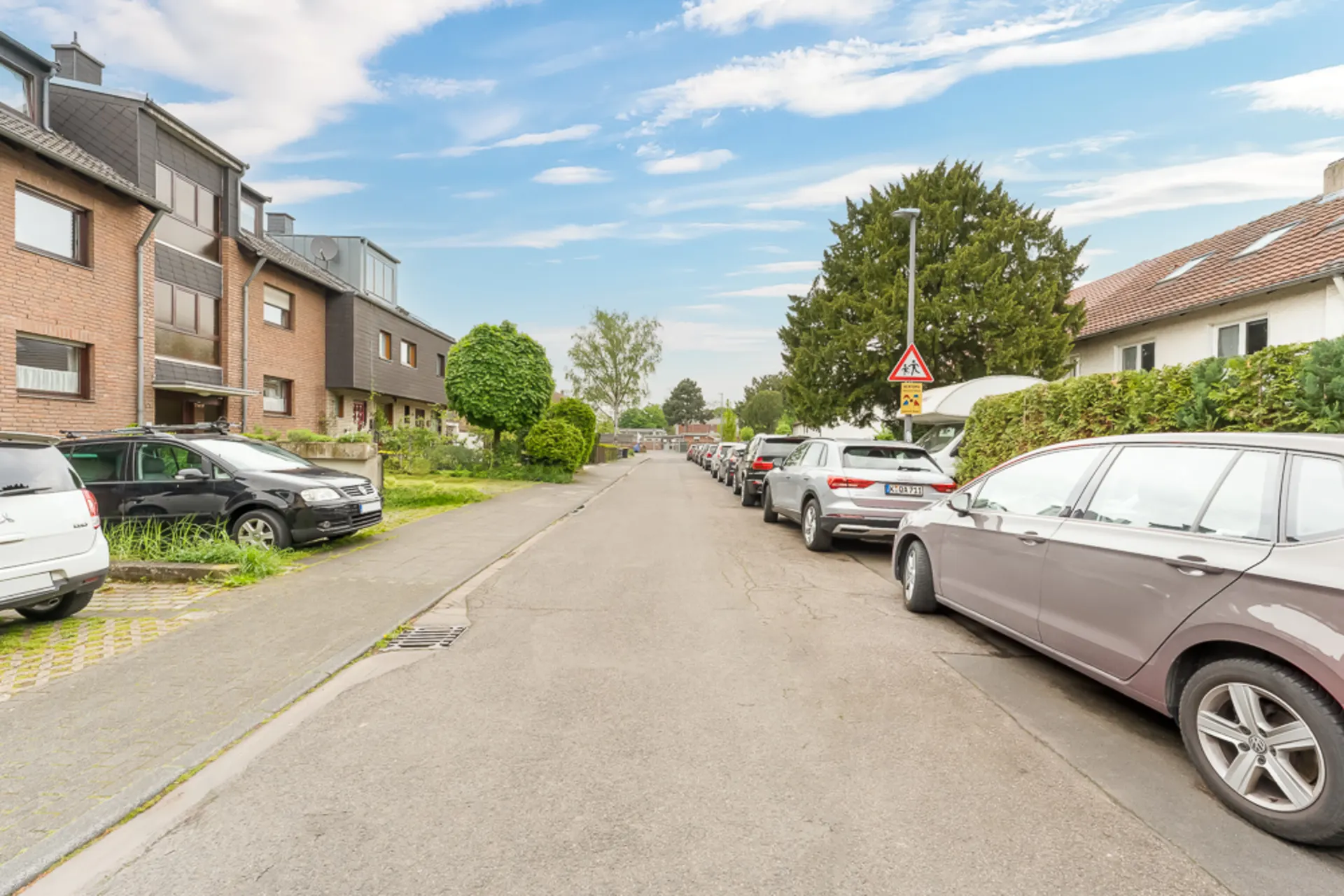 Ruhige Wohnstraße mit parkenden Autos, Häusern und Bäumen an beiden Seiten.