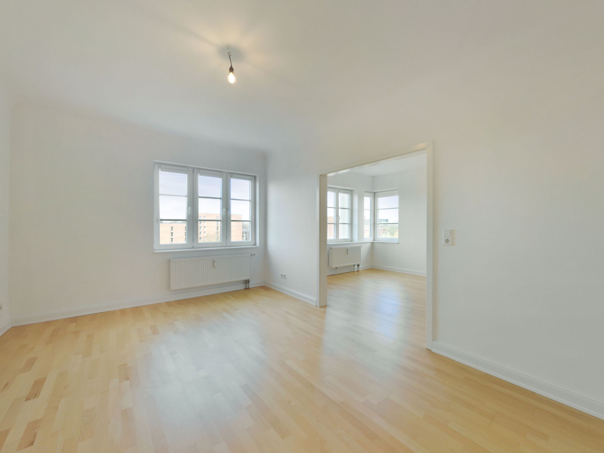 Helles, leeres Zimmer mit Parkettboden und großen Fenstern, verbunden mit einem weiteren Raum.