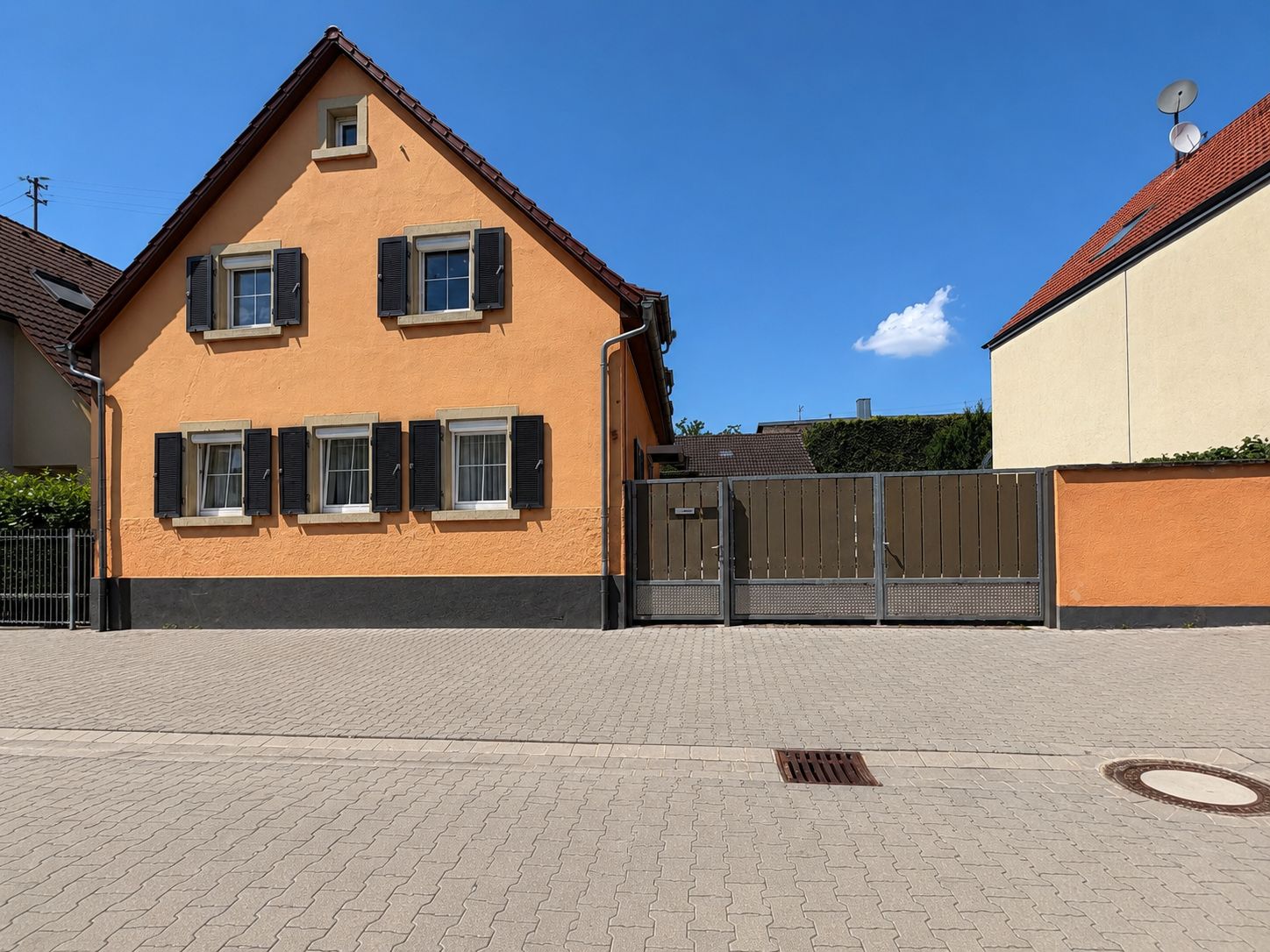 Orangefarbenes Wohnhaus mit schwarzen Fensterläden und großem Tor vor einem gepflasterten Hof.