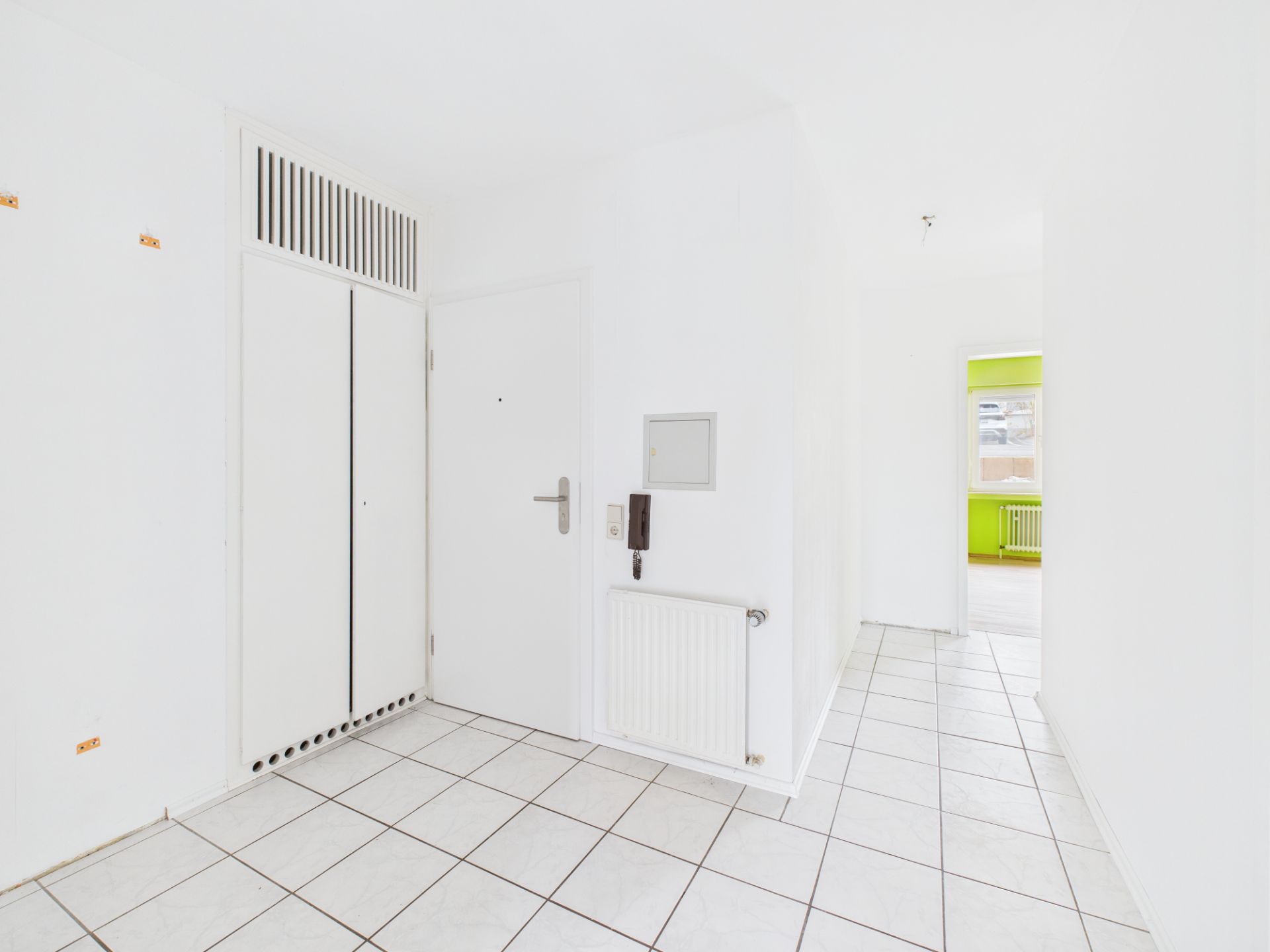 Heller Flur mit weißen Wänden, Fliesenboden und Türen, im Hintergrund ein Zimmer mit grünem Akzent.