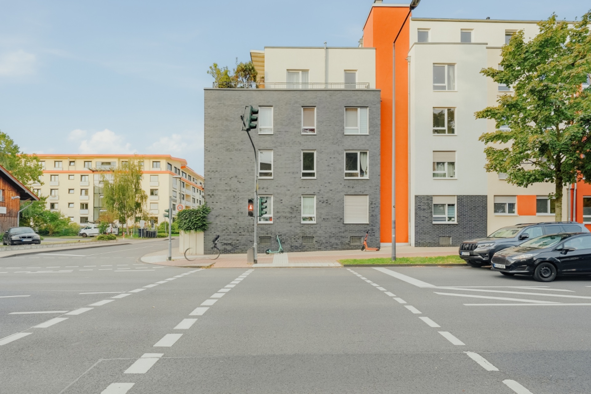 Mehrfamilienhaus mit grauer und orangefarbener Fassade an einer Straßenkreuzung, mehrere Autos parken davor.
