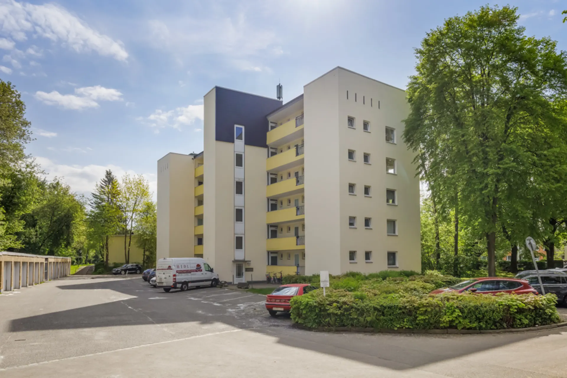 Mehrstöckiges Wohngebäude mit Balkonen, umgeben von Bäumen und geparkten Autos.