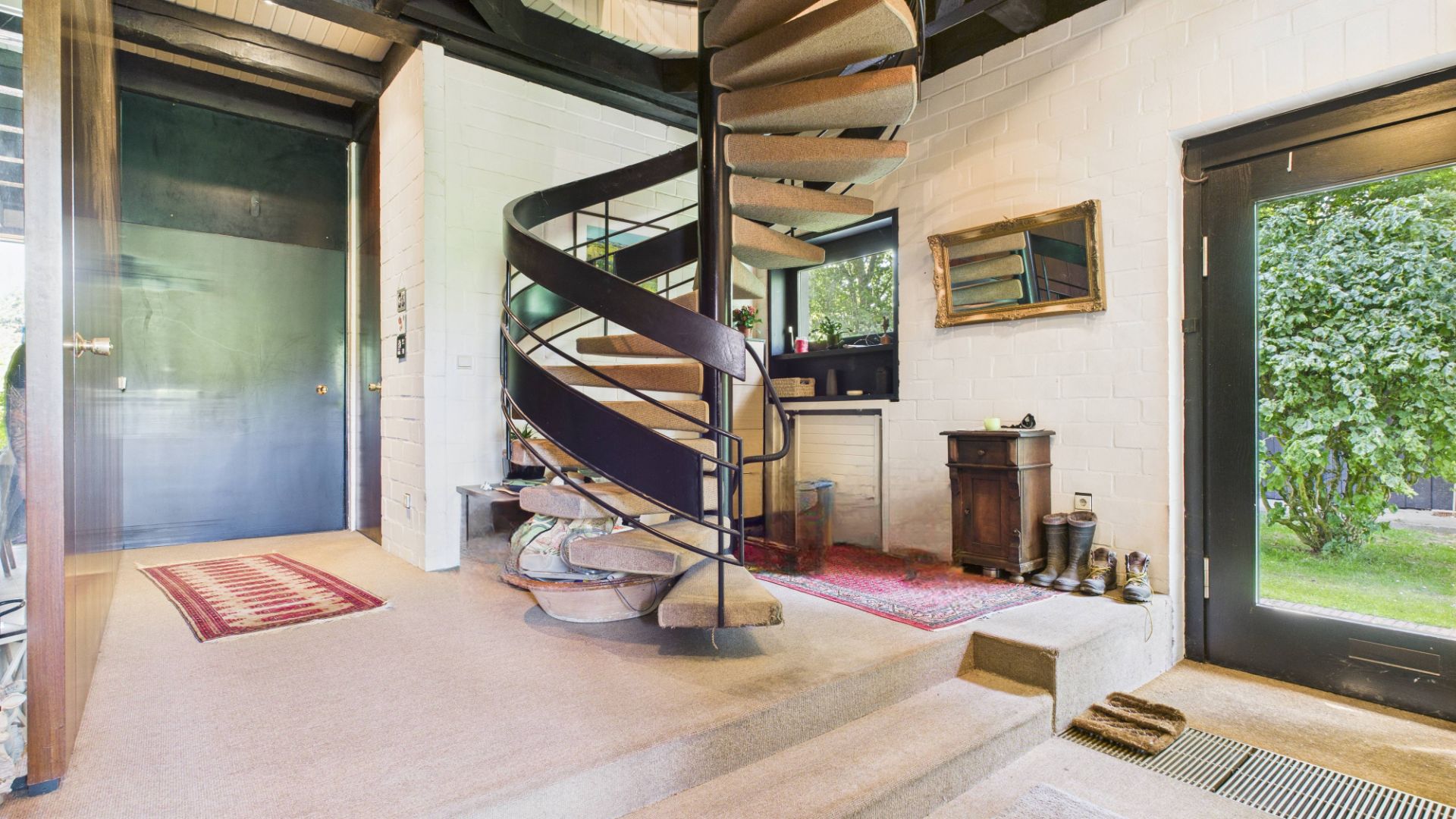 Heller Wohnraum mit Wendeltreppe, großem Fenster, Teppichen und kleinem Tisch mit Spiegel.
