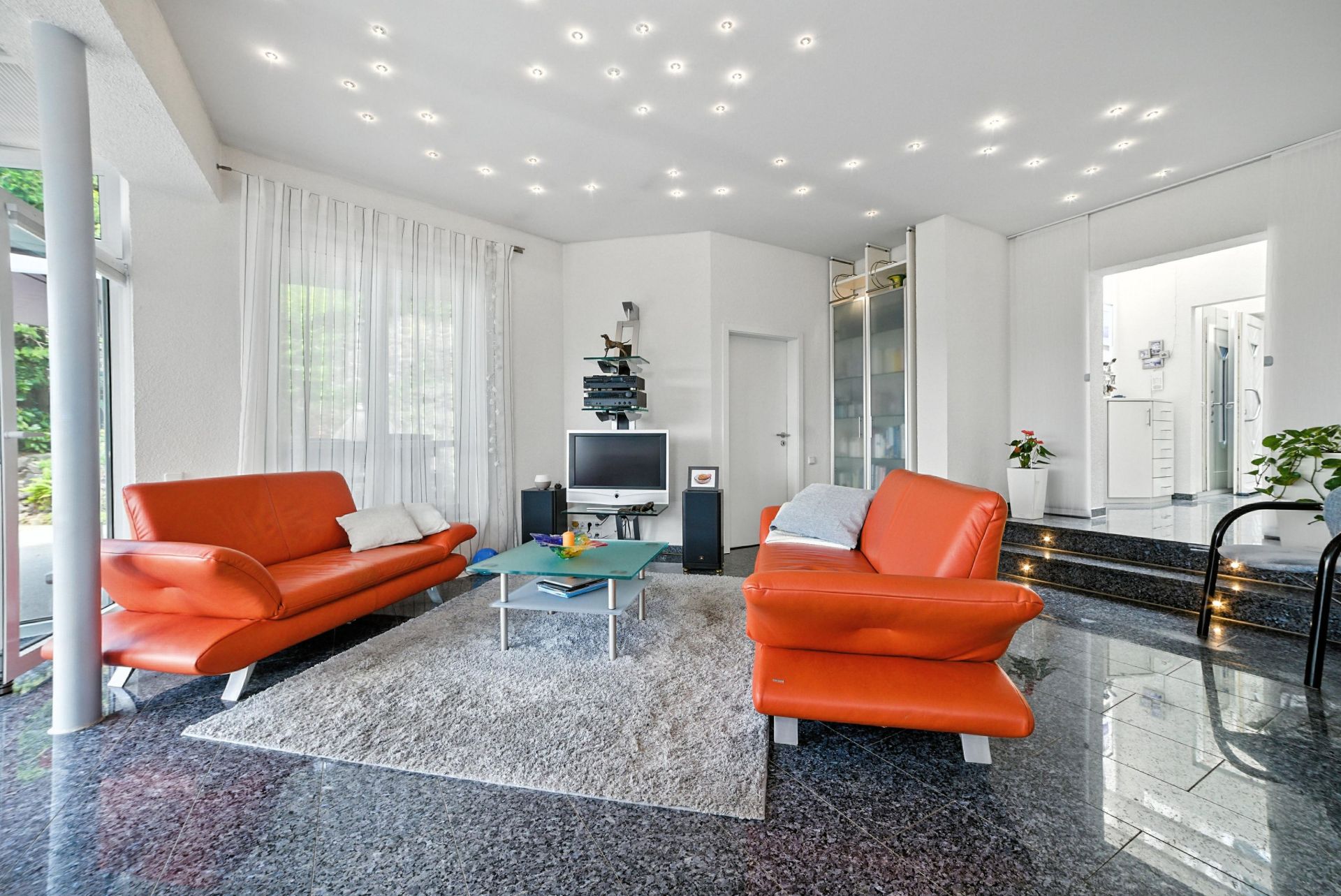 Modernes Wohnzimmer mit zwei orangefarbenen Sofas, Glastisch und Fernseher auf glänzendem Boden.