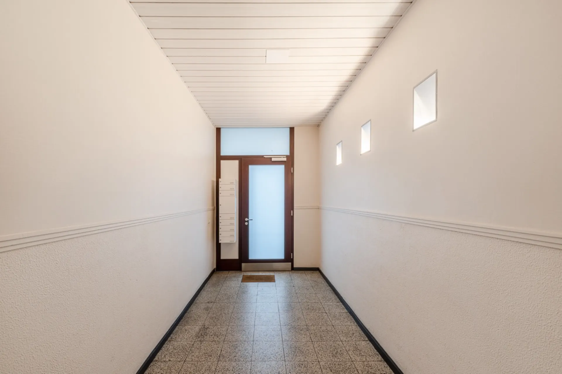 Heller Flur mit Fliesenboden, Briefkästen und Tür mit Milchglas in einem Wohnhaus.