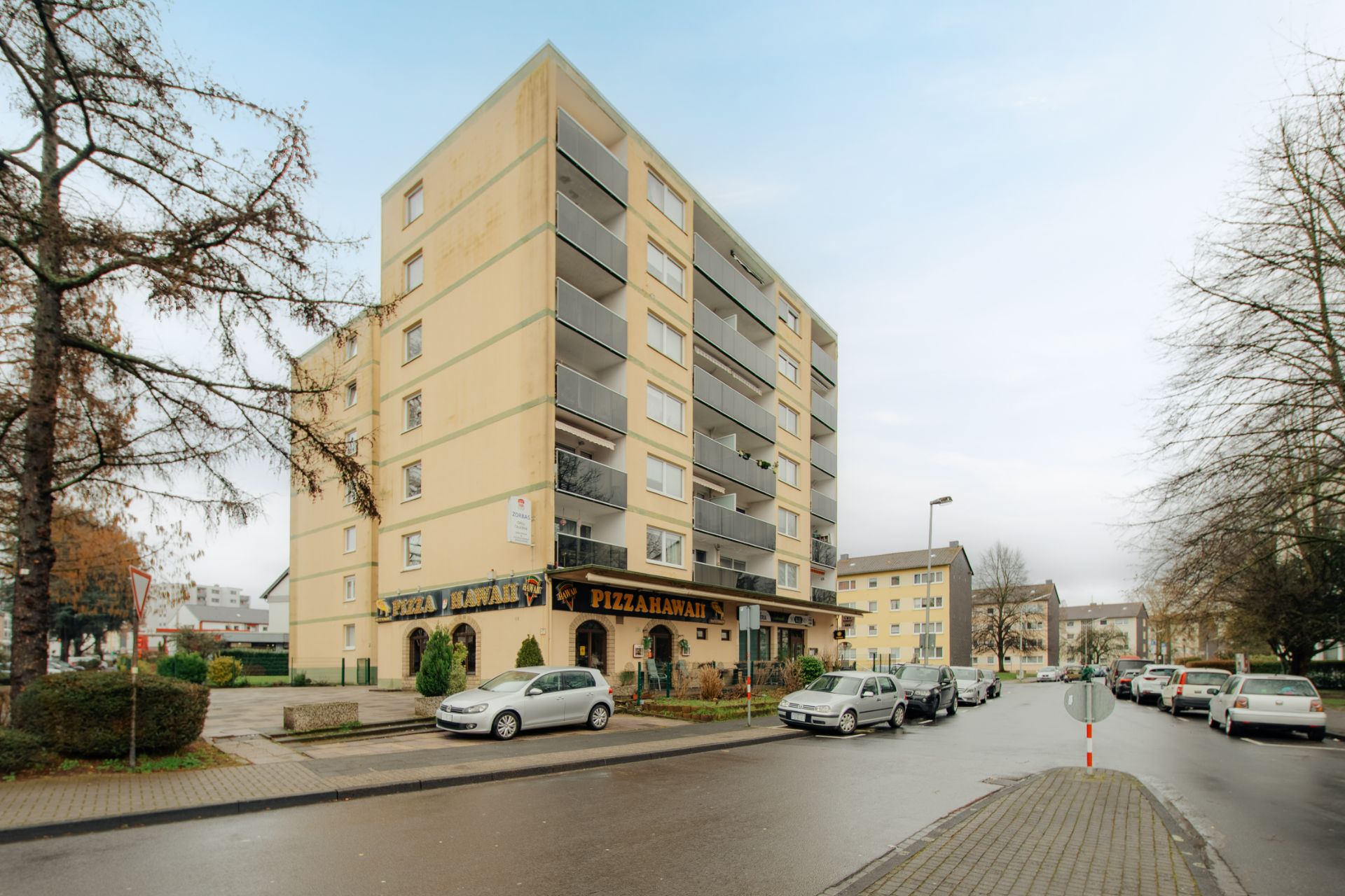 Mehrfamilienhaus mit mehreren Balkonen und parkenden Autos auf der Straße.