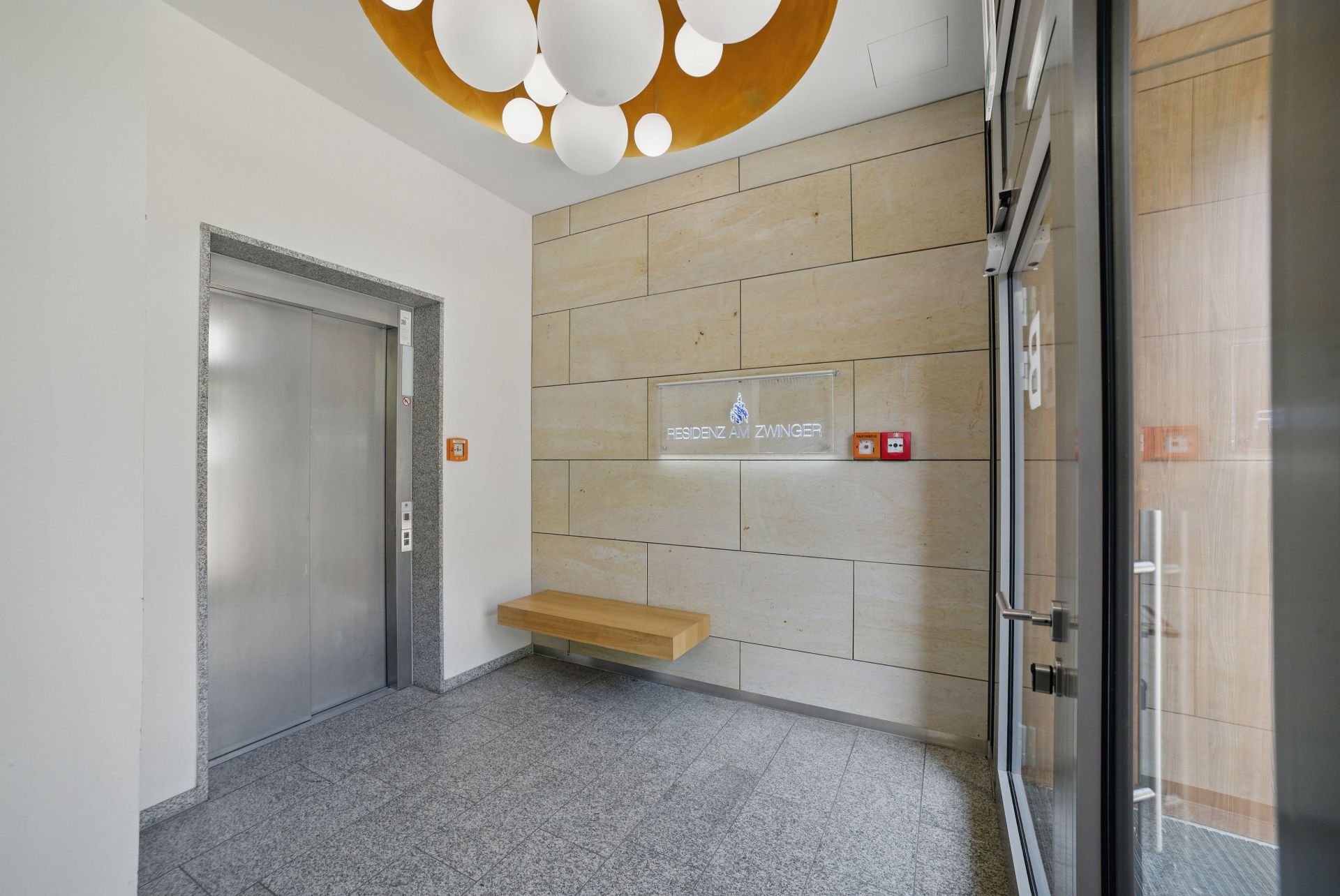 Heller Flur mit Aufzug, Holzbank an der Wand und runder Deckenleuchte.