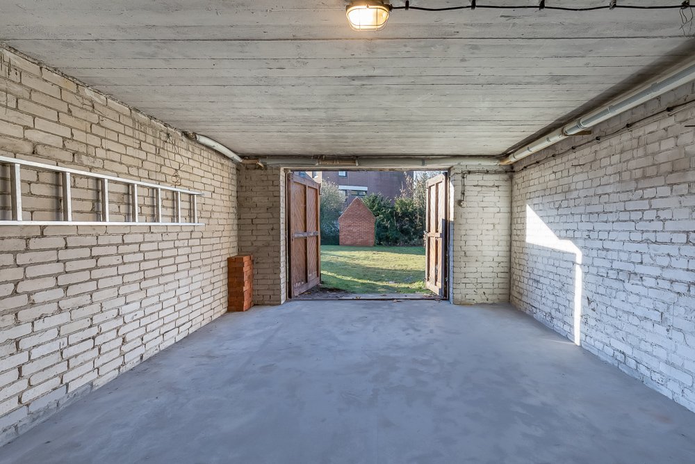 Leere Garage mit weißen Backsteinwänden, geöffneter Holztür und Blick auf einen Garten.