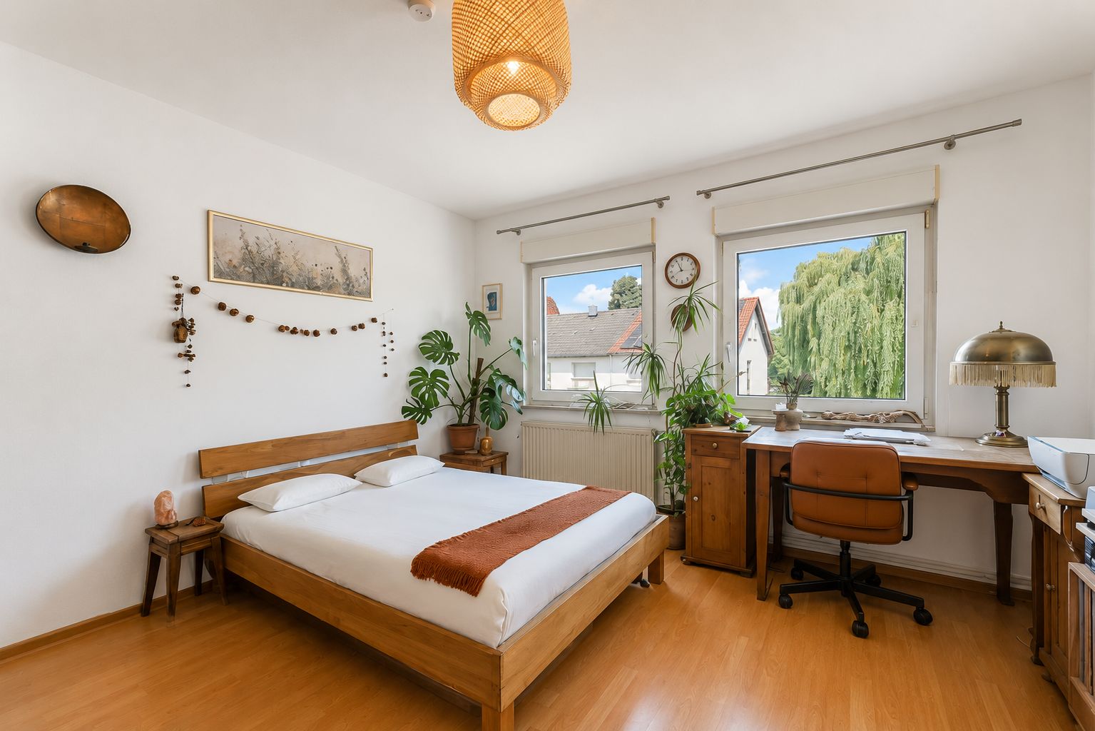 Helles Schlafzimmer mit Holzbett, Schreibtisch, Stuhl, Zimmerpflanzen und zwei Fenstern.