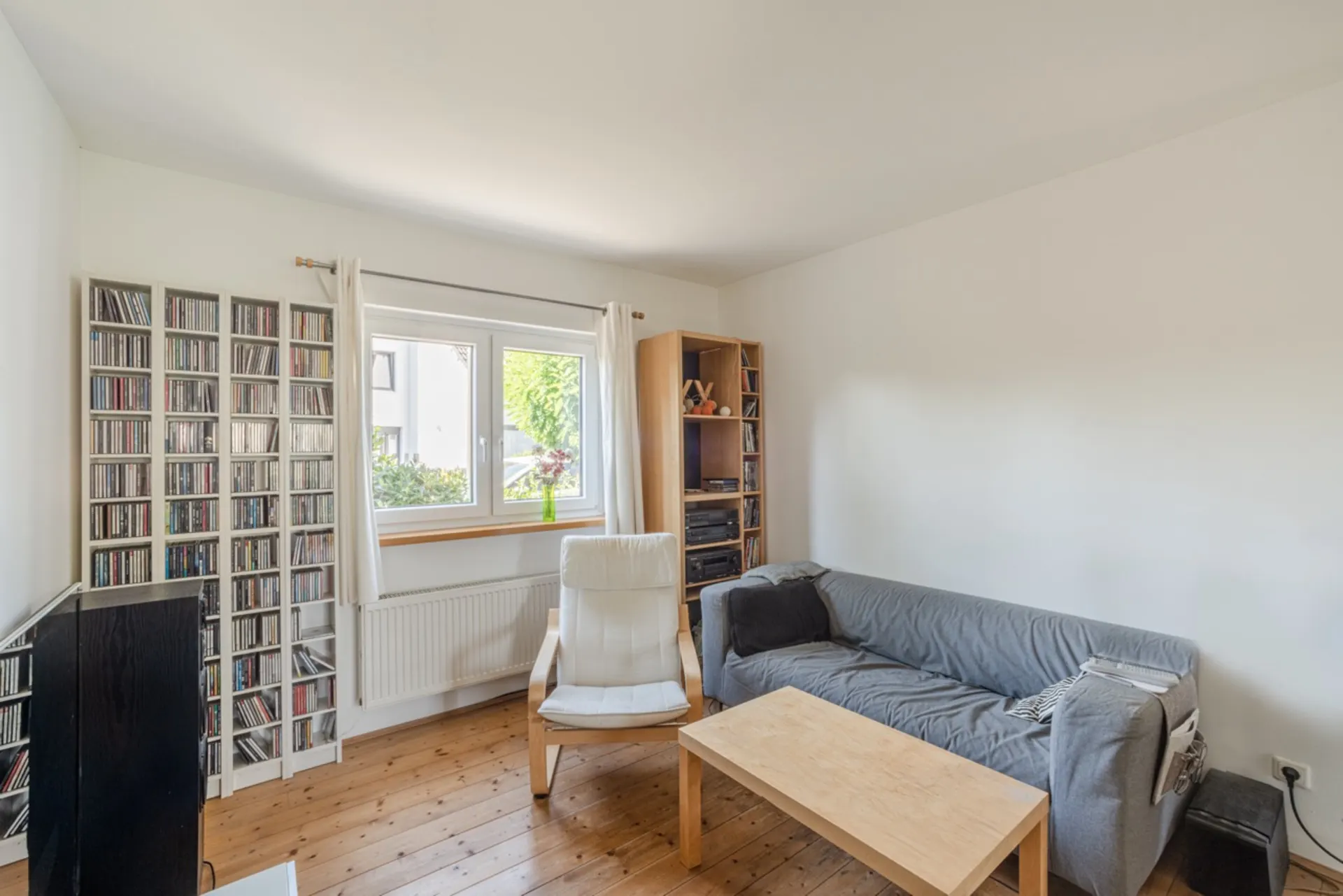 Wohnzimmer mit grauem Sofa, Sessel, Holztisch, CD-Regalen und Fenster mit Blick nach draußen.