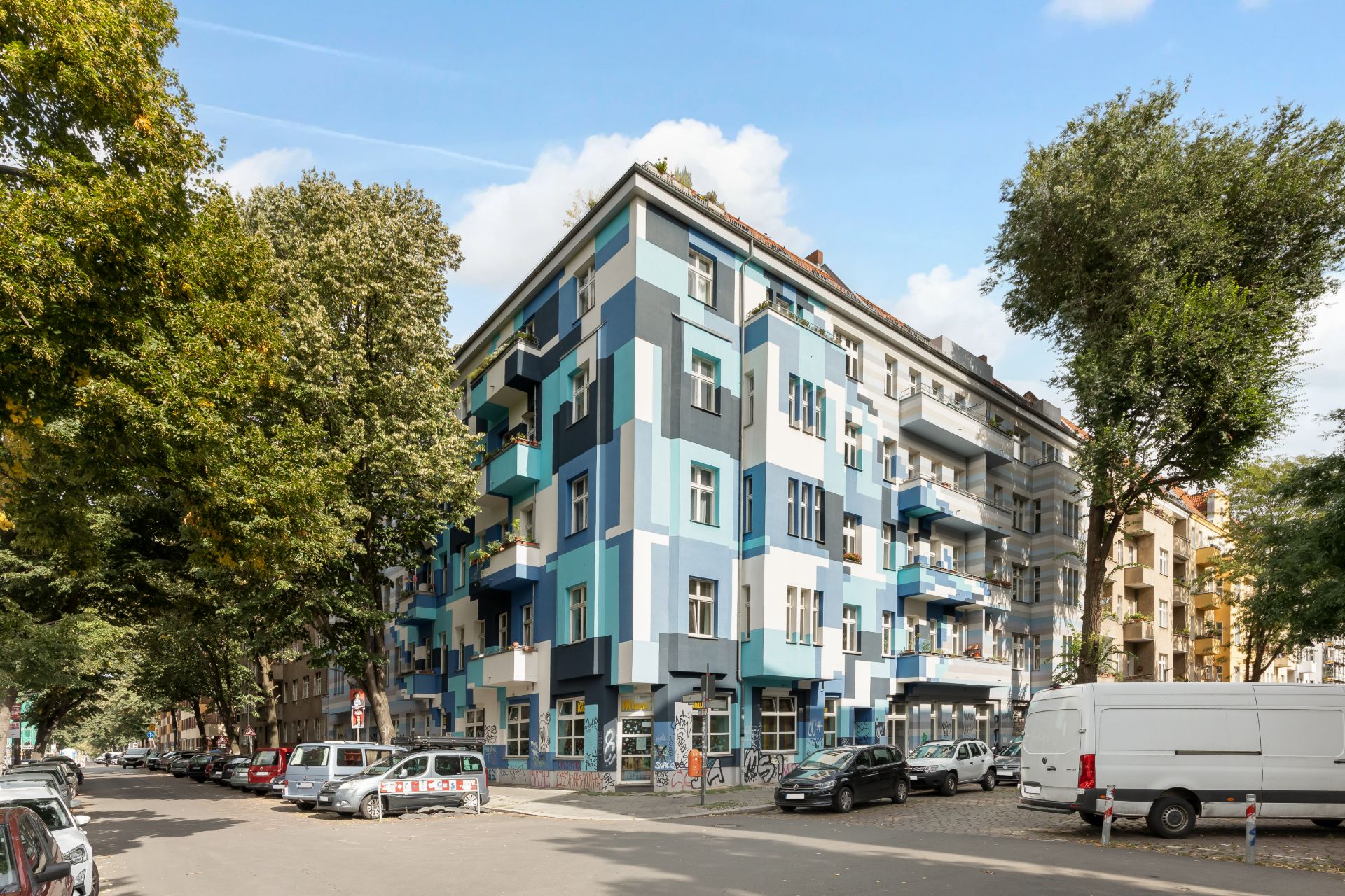 Eckwohnhaus mit blauer Fassade und Balkonen, umgeben von Bäumen und parkenden Autos.