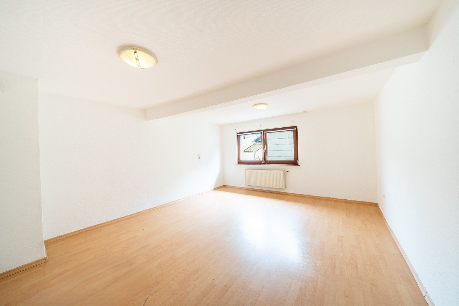 Helles, leeres Zimmer mit Holzfußboden, weiß gestrichenen Wänden und einem Fenster mit Heizkörper darunter.