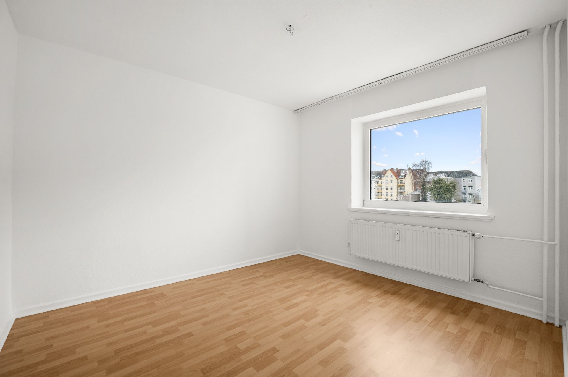 Leeres Zimmer mit weißen Wänden, Holzfußboden und großem Fenster mit Blick nach draußen.