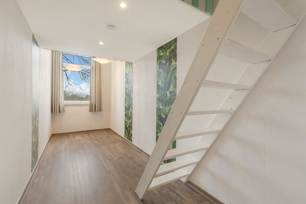 Helles Zimmer mit Holzfußboden, großem Fenster, Vorhängen und weißer offener Treppe.