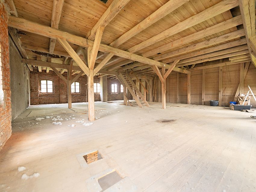 Leerer Dachboden mit sichtbaren Holzträgern, Holzboden und Fenstern, etwas Bauschutt am Boden.