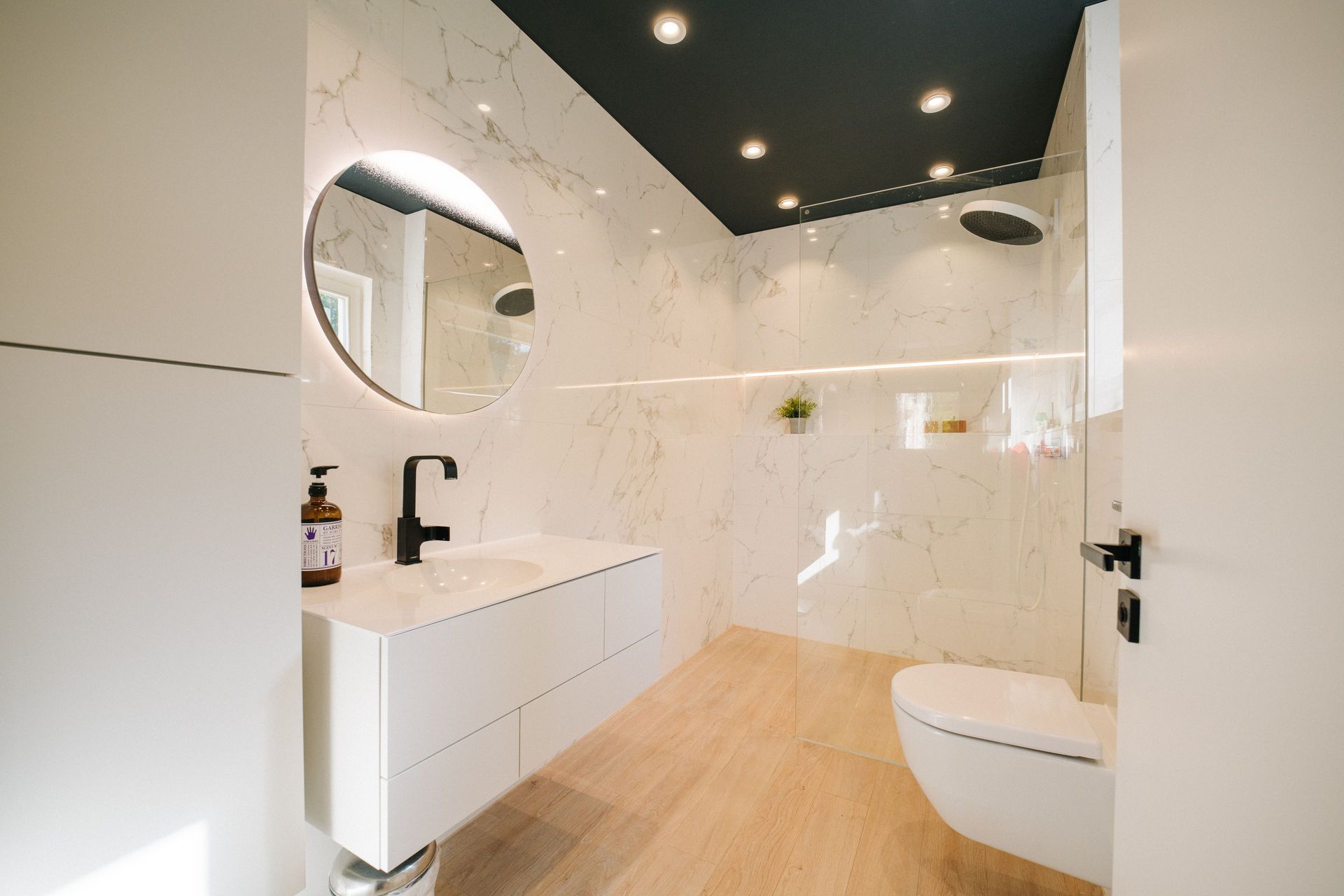 Modernes Bad mit weißem Waschbecken, rundem Spiegel, WC und großer Dusche auf Holzfußboden.