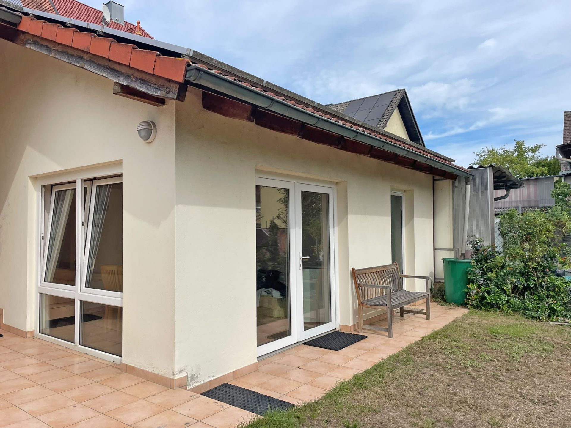 Erdgeschossiges Haus mit Terrasse, weißen Fenstern und einer Bank, daneben etwas Grünfläche.