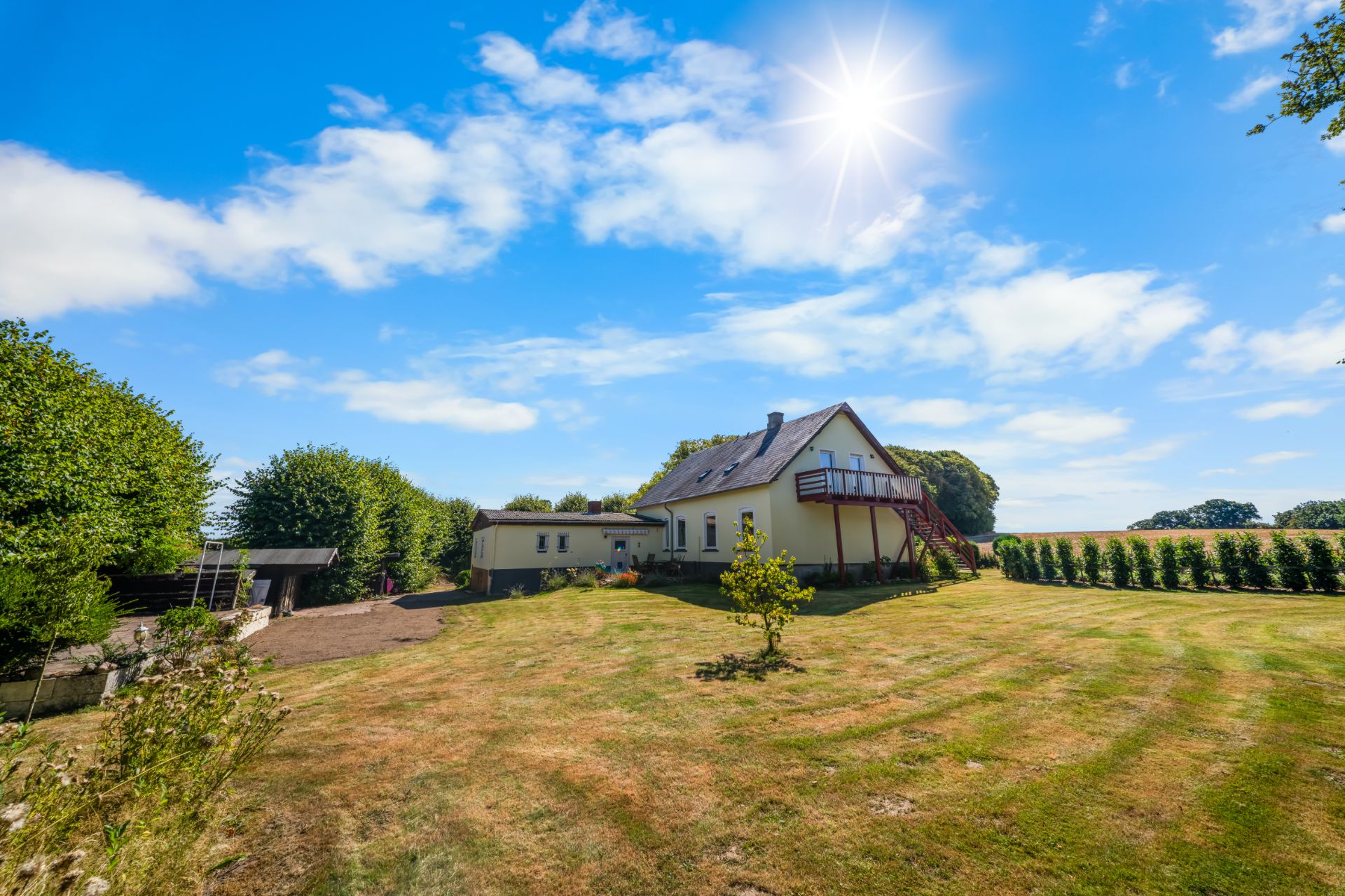 Haus mit großem Garten, Rasenfläche und vereinzelten Bäumen unter blauem Himmel mit Sonne.