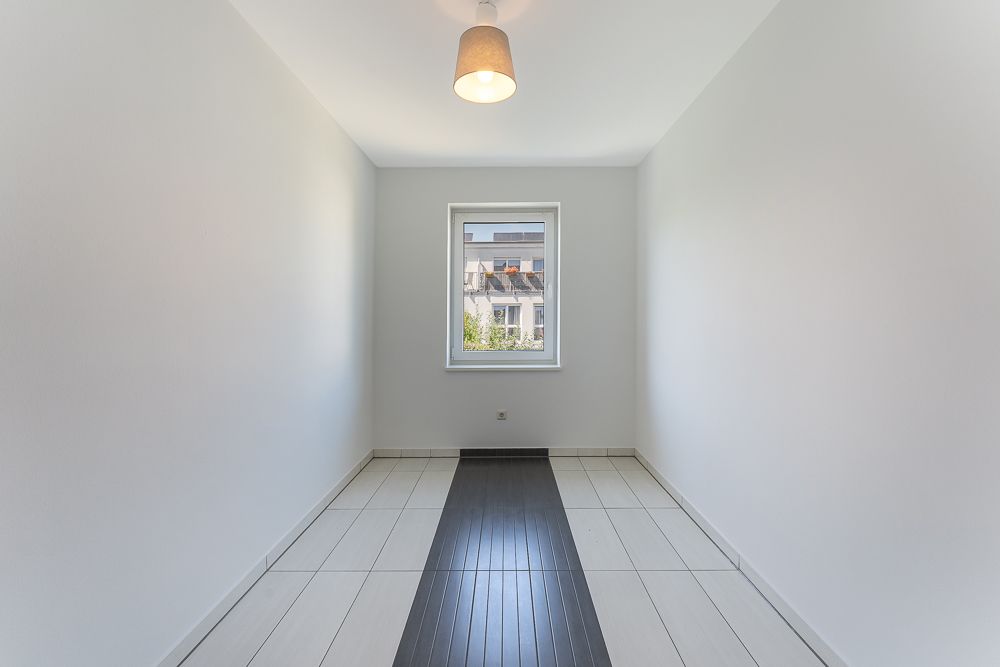 Schmaler, leerer Raum mit Fliesenboden, weißer Wand und Fenster am Ende.