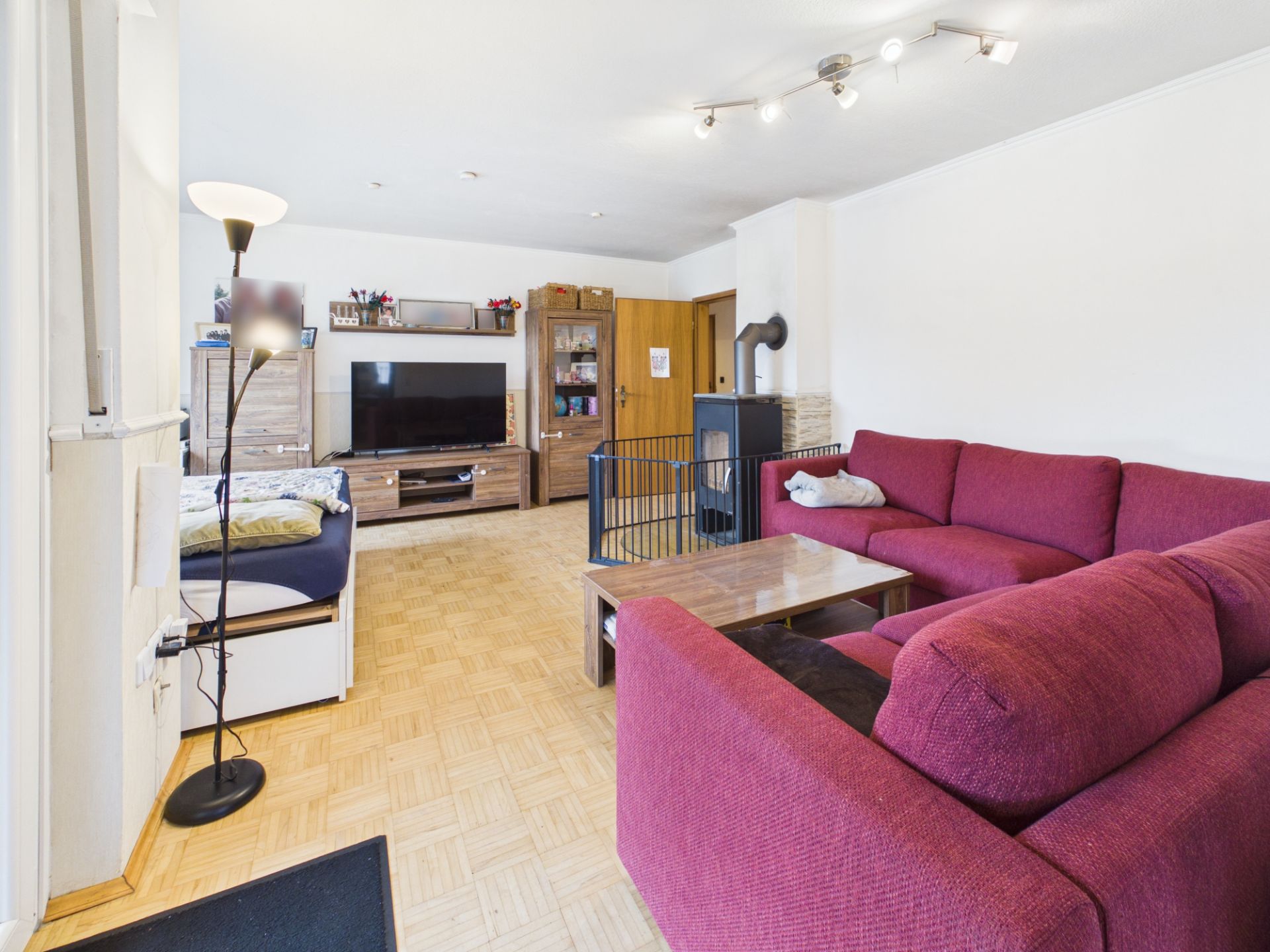Wohnzimmer mit rotem Ecksofa, Couchtisch, TV, Schrank, Stehlampe und Holzofen.