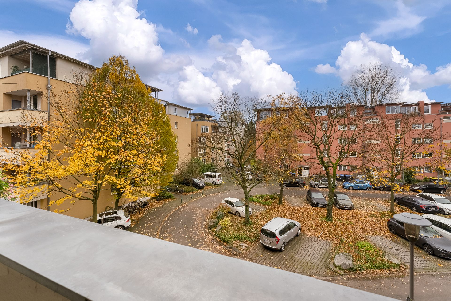 Blick auf einen Parkplatz mit Autos, Bäumen mit Herbstlaub und Wohngebäuden im Hintergrund.