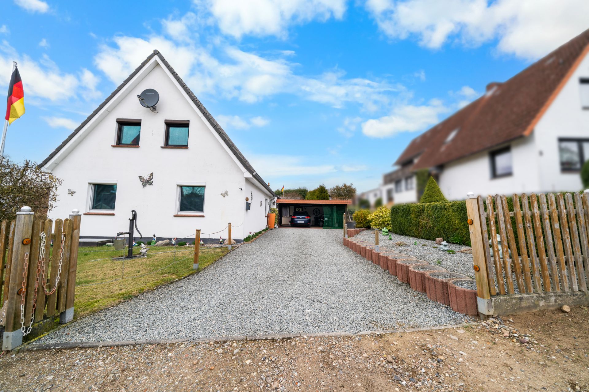 Einfamilienhaus mit Spitzdach, Vorgarten, Kiesauffahrt und Carport, deutsche Flagge links.