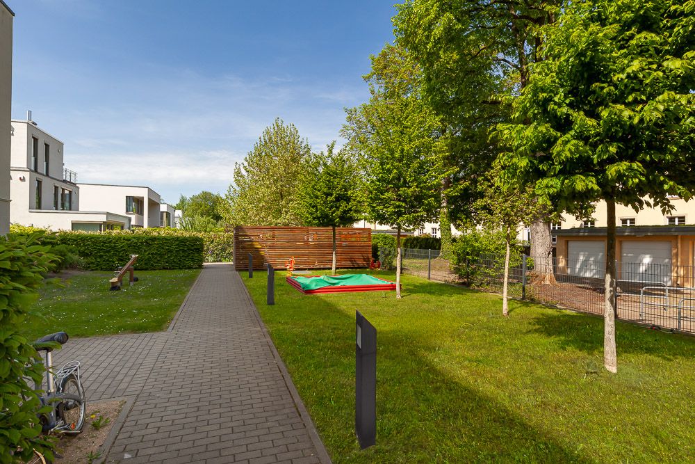 Gepflasterter Weg im Garten, Sandkasten, Bäume und moderne Wohngebäude im Hintergrund.