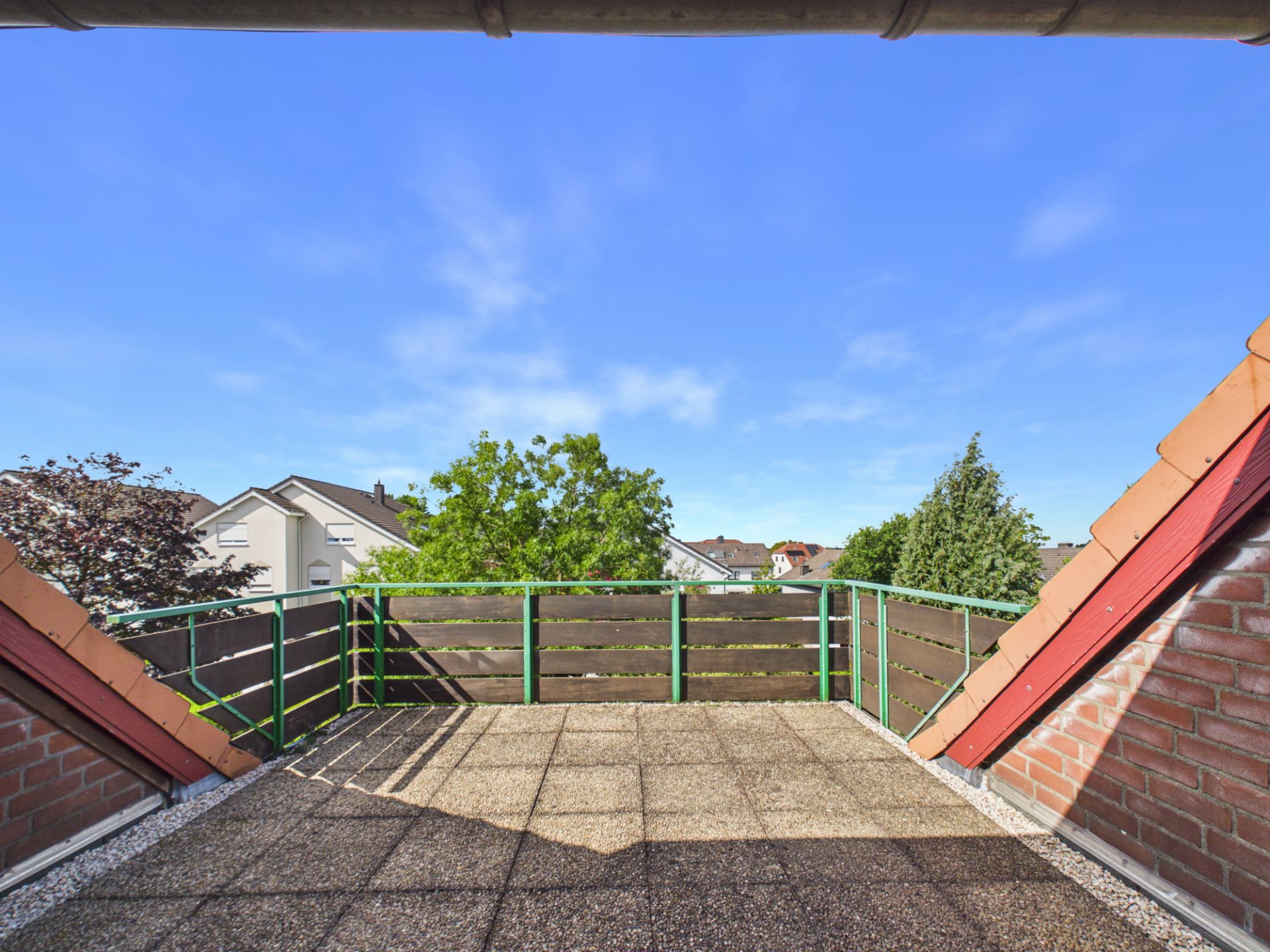 Dachterrasse mit Fliesenboden, Geländer und Blick auf Bäume und Nachbarhäuser bei blauem Himmel.