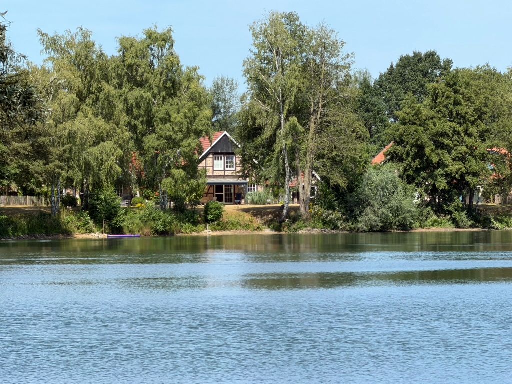 Haus mit rotem Dach am Seeufer, umgeben von Bäumen, bei sonnigem Wetter.