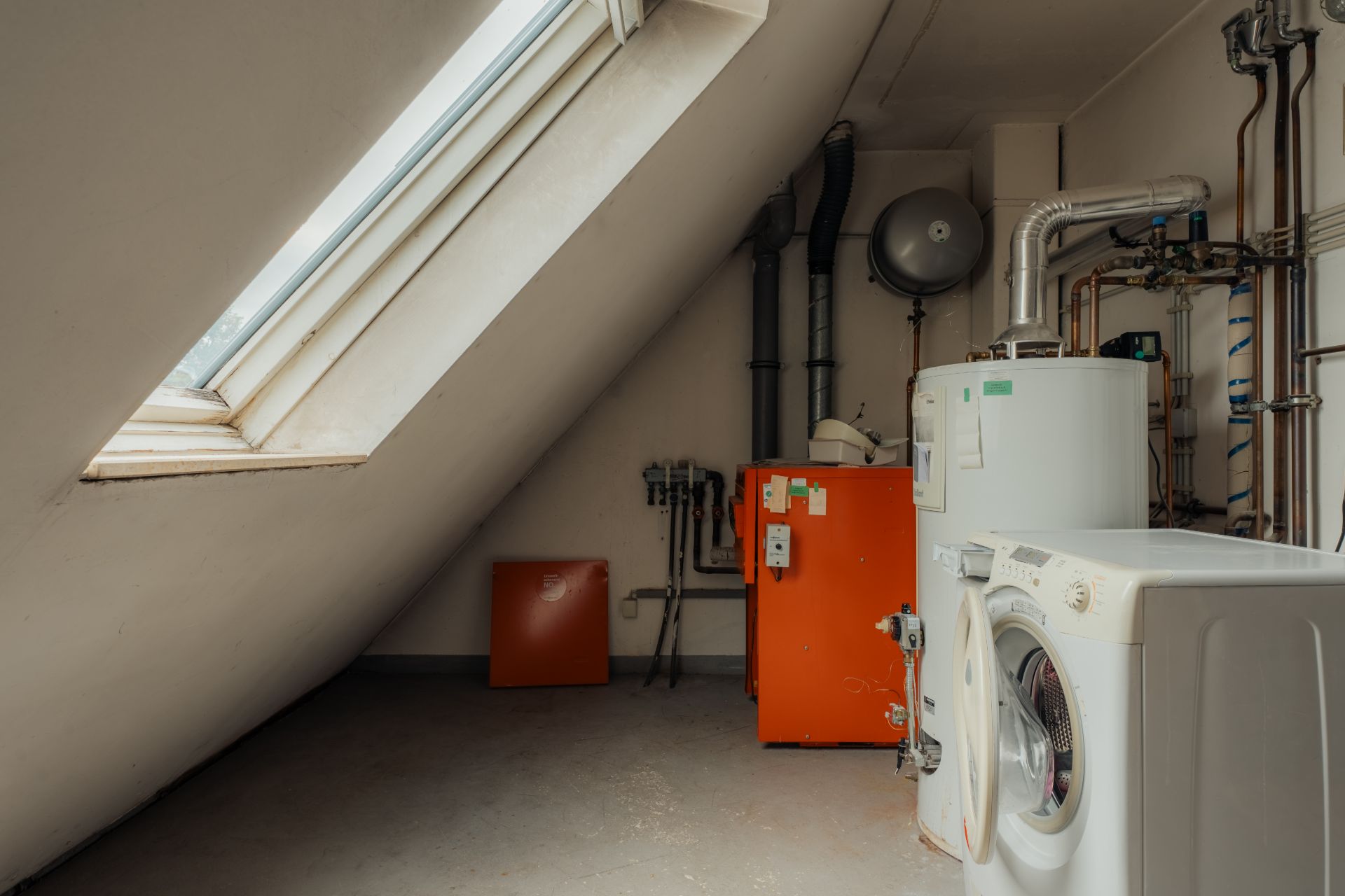 Hauswirtschaftsraum mit Waschmaschine, Heizkessel und Rohrleitungen unter einem Dachfenster.