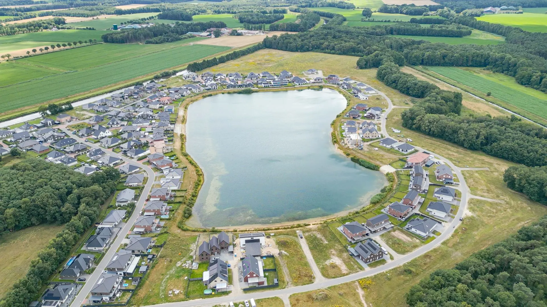 Luftaufnahme eines Wohngebiets mit Häusern rund um einen zentralen See, umgeben von Feldern und Wald.