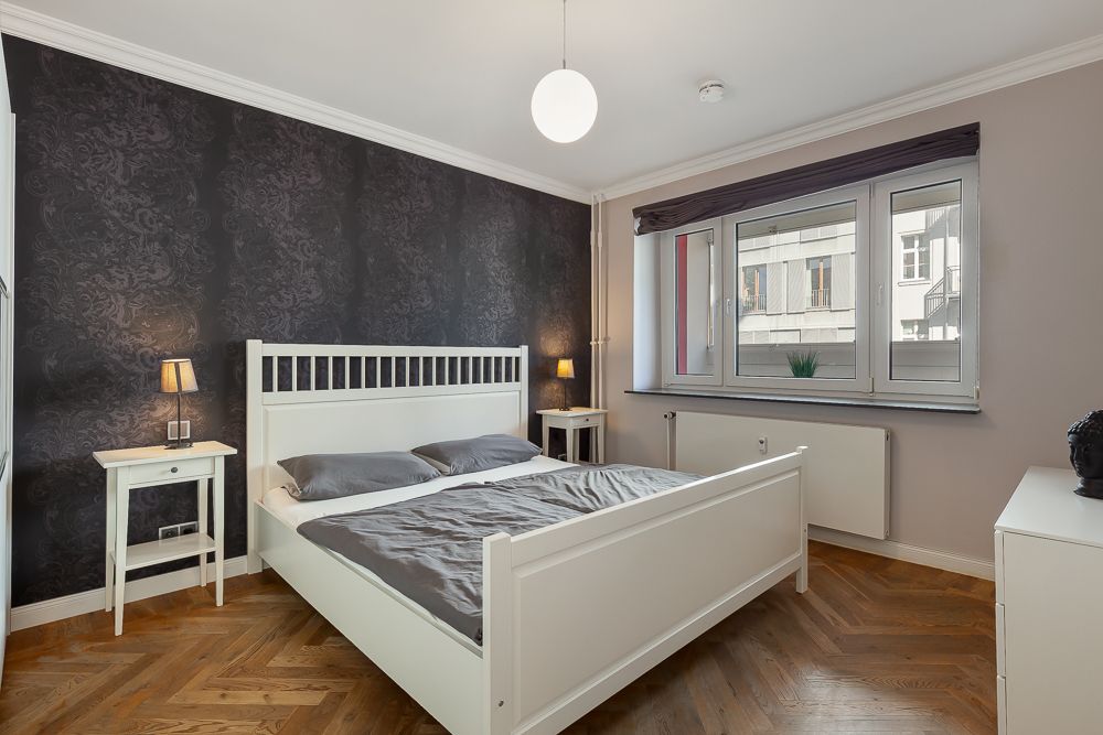 Schlafzimmer mit weißem Bett, zwei Nachttischen, zwei Lampen und großem Fenster.