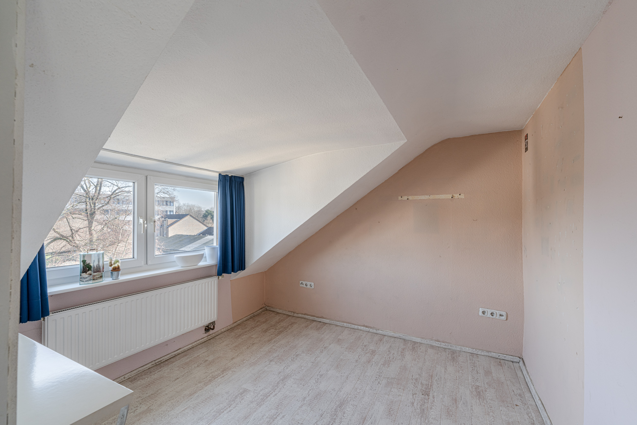 Leeres Zimmer mit Dachschräge, großem Fenster, blauen Vorhängen und hellem Boden.