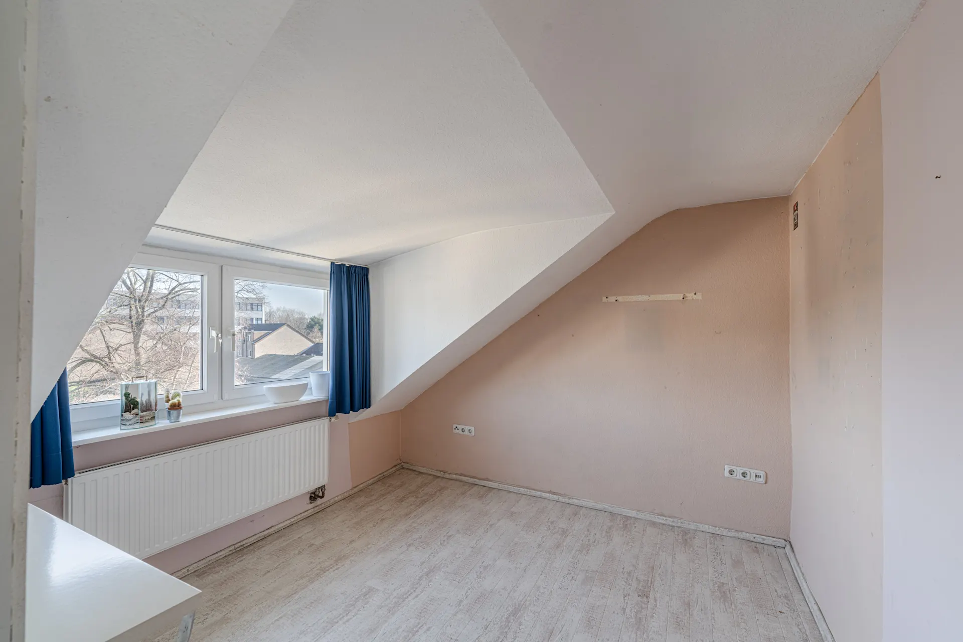 Leeres Zimmer mit Dachschräge, großem Fenster, blauen Vorhängen und hellem Boden.