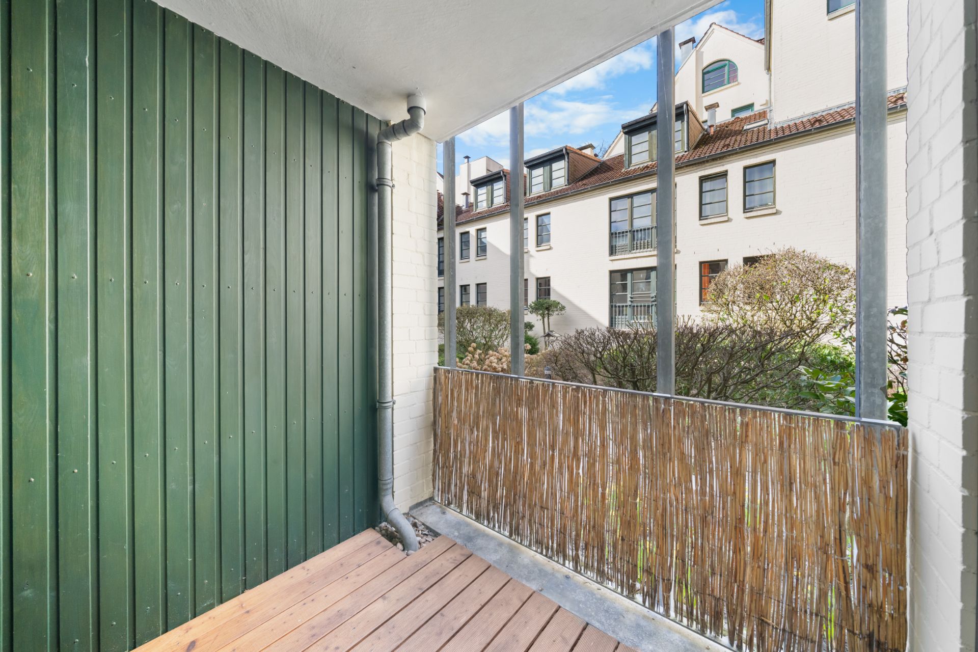 Überdachter Balkon mit Holzboden, grünem Sichtschutz und Blick auf Wohnhäuser und Sträucher.
