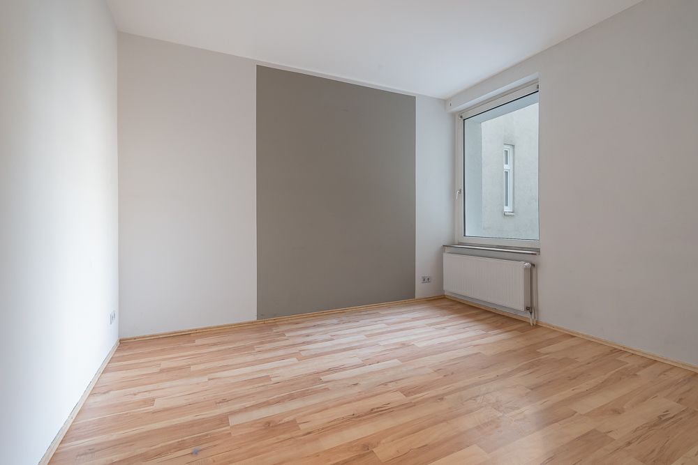 Leerer Raum mit Holzboden, grauer Akzentwand und großem Fenster mit Heizkörper darunter.