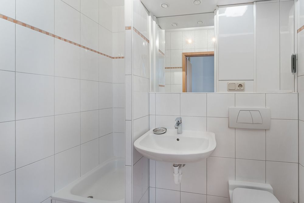 Kleines, weiß gefliestes Badezimmer mit Waschbecken, Toilette und Dusche.