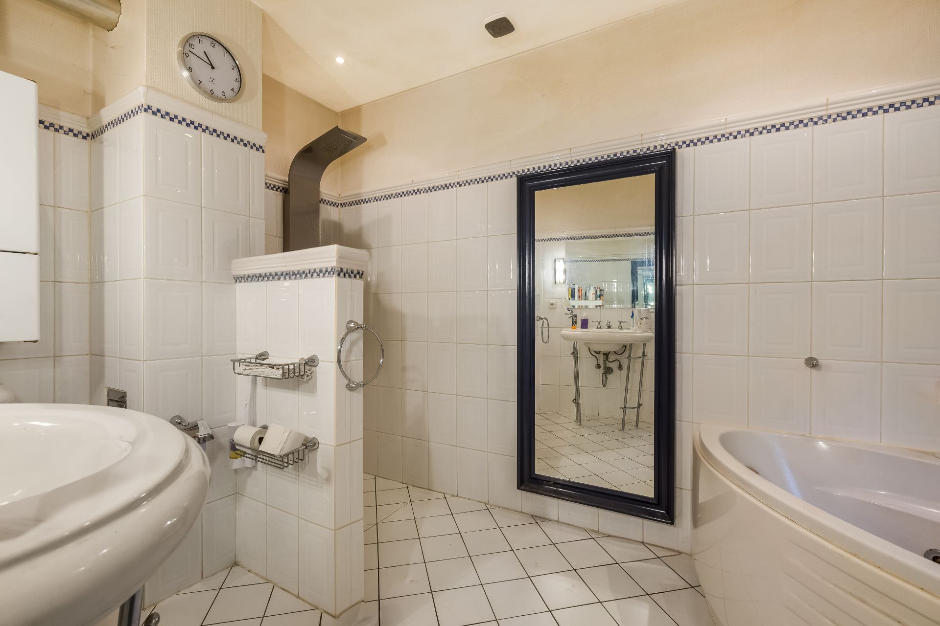 Helles Badezimmer mit Badewanne, großem Spiegel, Uhr und weiß gefliesten Wänden.