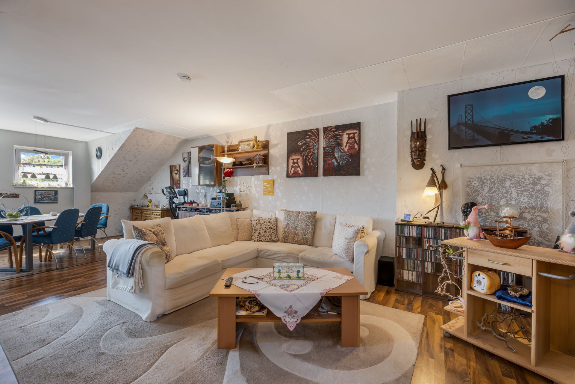 Wohnzimmer mit heller Eckcouch, Couchtisch, Regal und Essbereich im Hintergrund.