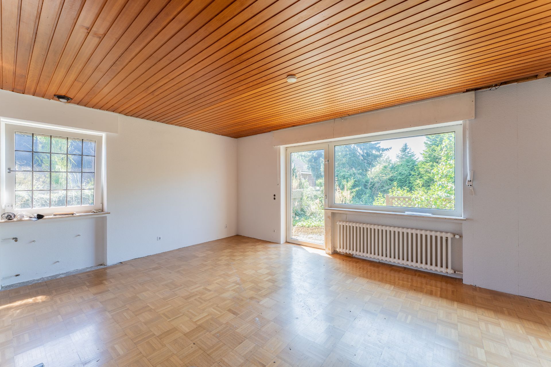 Leeres Zimmer mit Holzdecke, Parkettboden und großen Fenstern mit Blick ins Grüne.