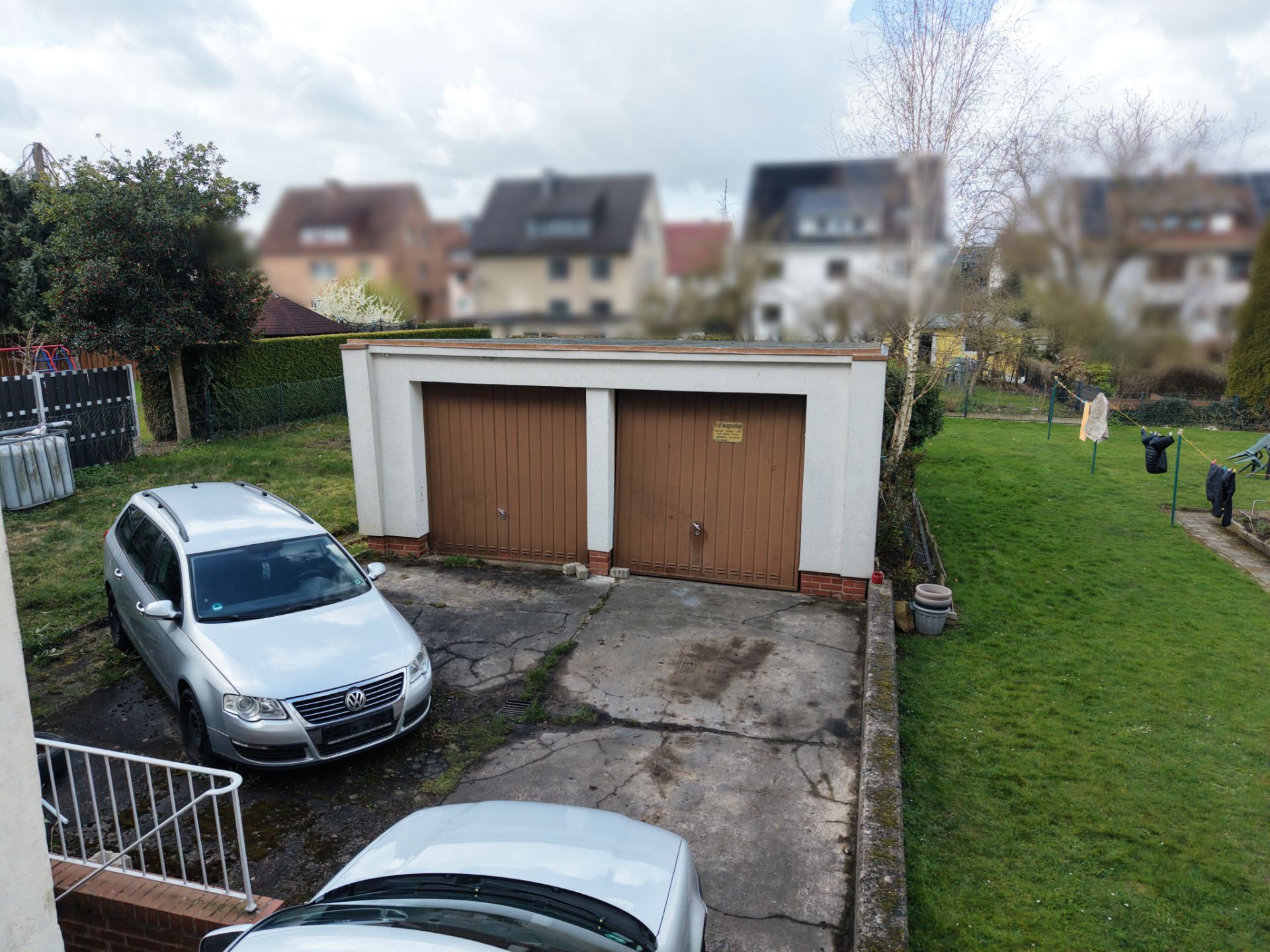 Zwei Garagen mit braunen Toren, zwei parkende Autos und ein großer Garten im Hintergrund.