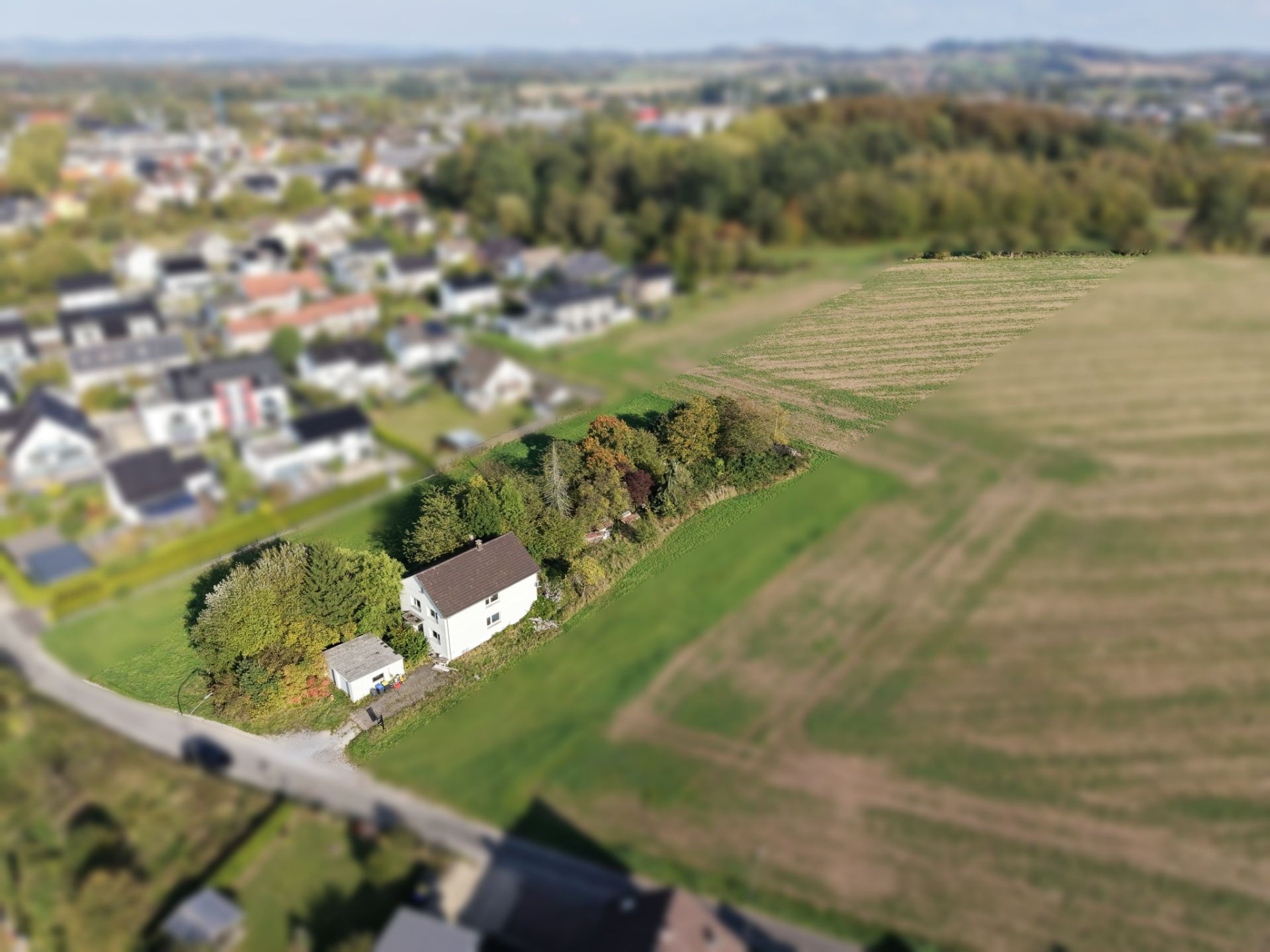Luftaufnahme eines weißen Hauses mit Garten am Rand eines Feldes, nahe einer Wohnsiedlung.