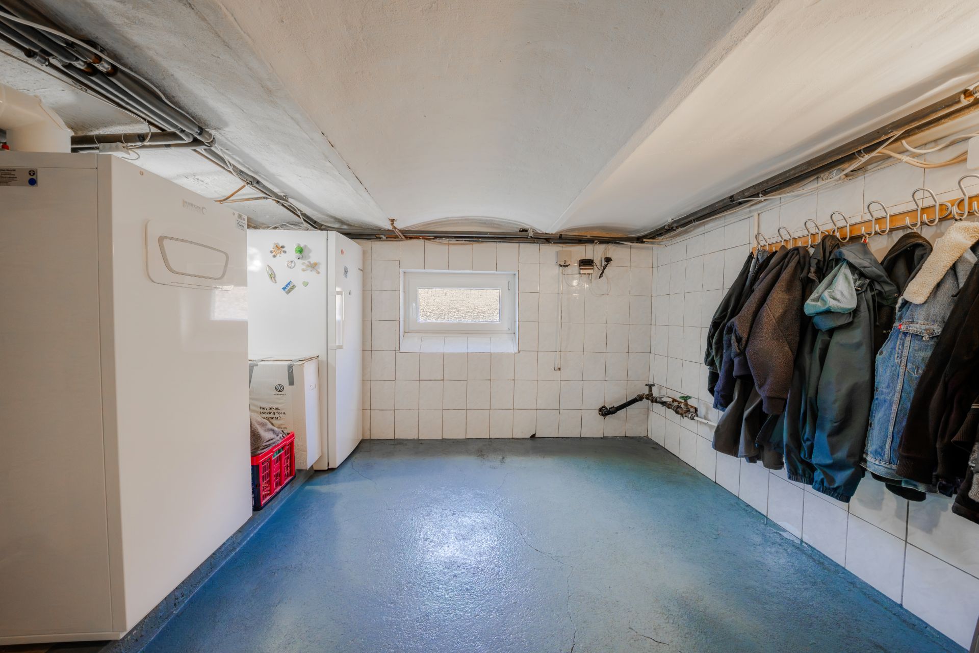 Kellerraum mit gefliestem Boden, Fenster, Kühlgeräten und Jacken an der Wand.